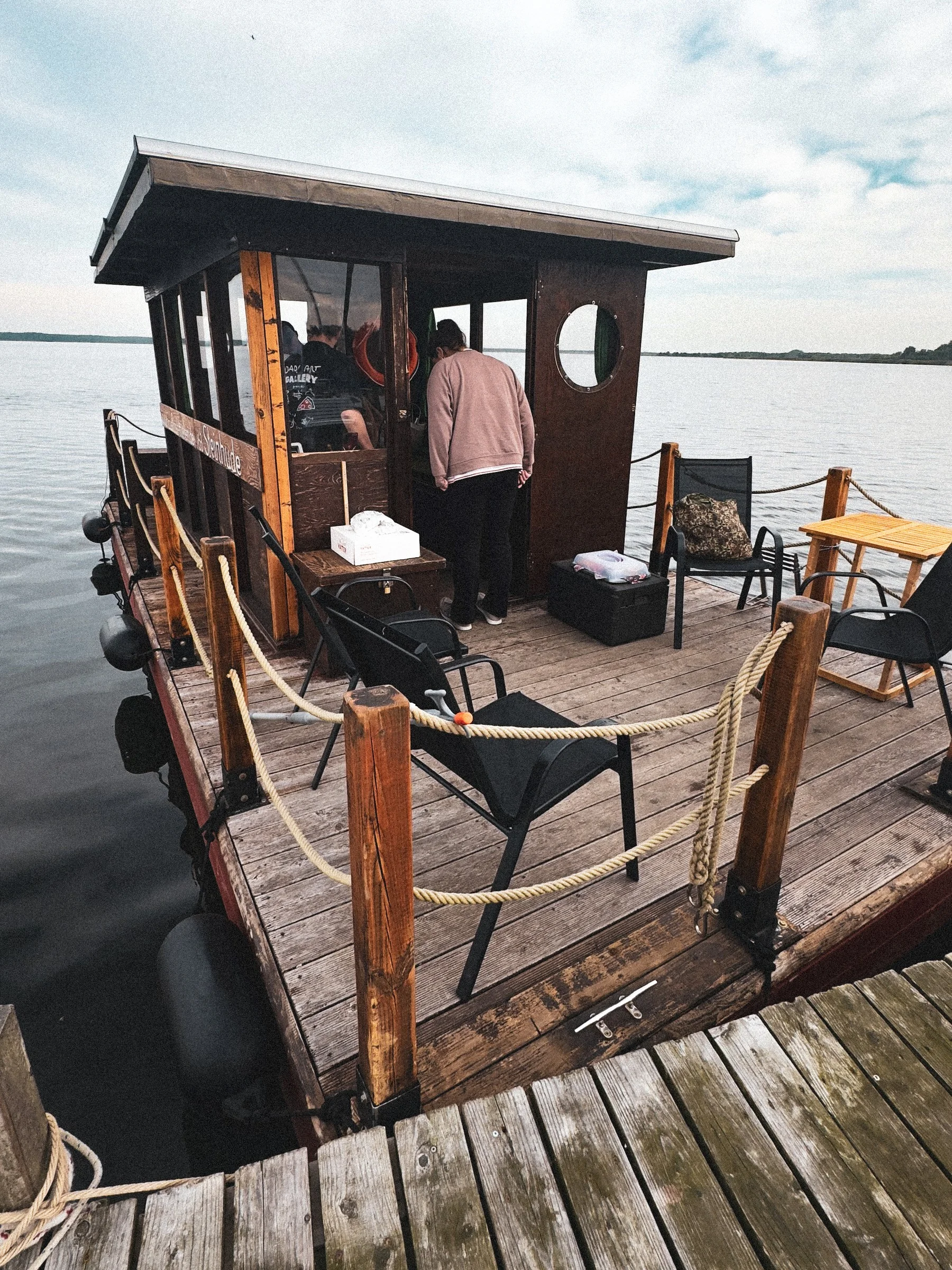 Jemand steht an der Ausgabe einer hölzernen Hütte auf einem Steg am Wasser, davor Stühle und Tische