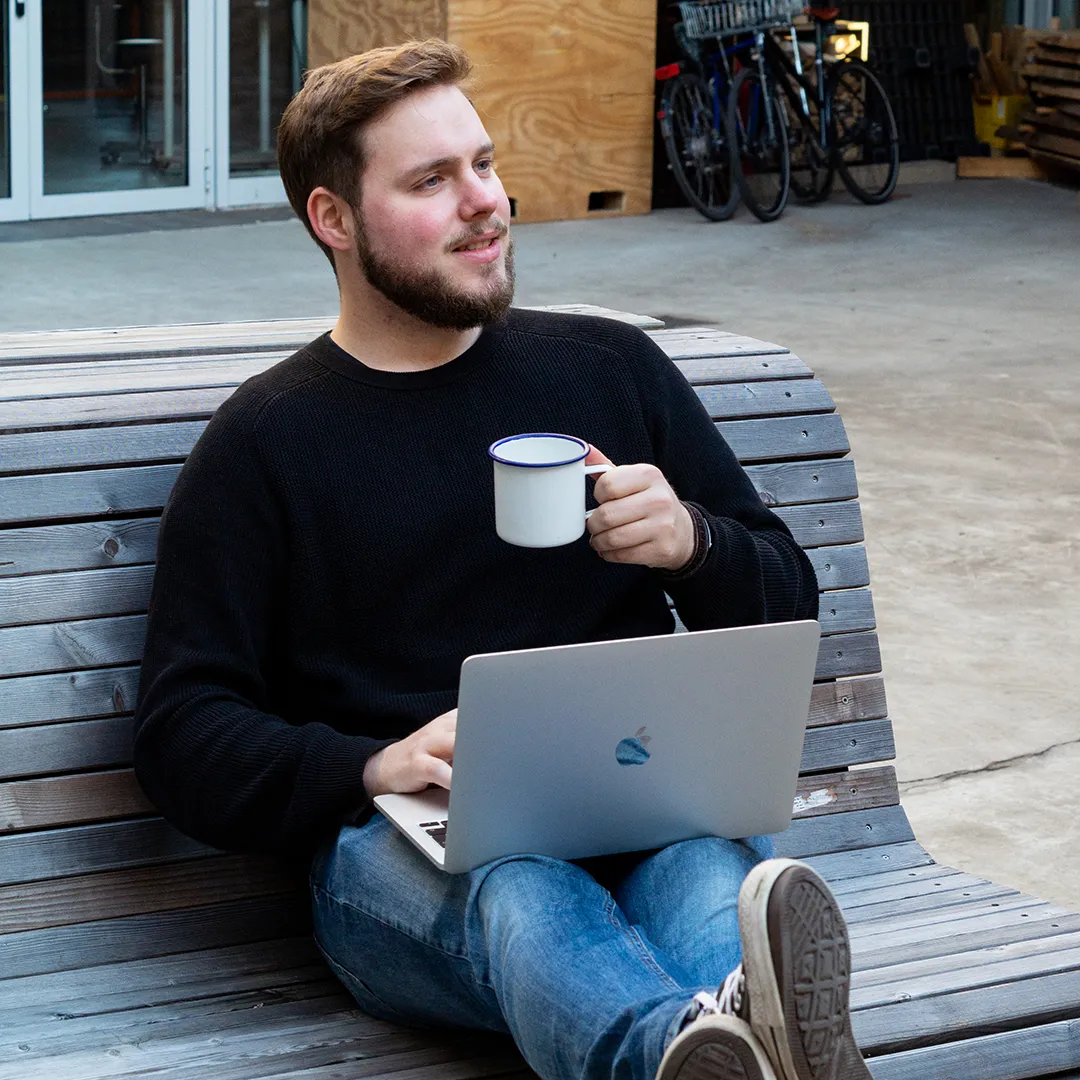 Tim sitzt mit dem Laptop und einem Emaillebecher auf der Holzbank vor dem Büro